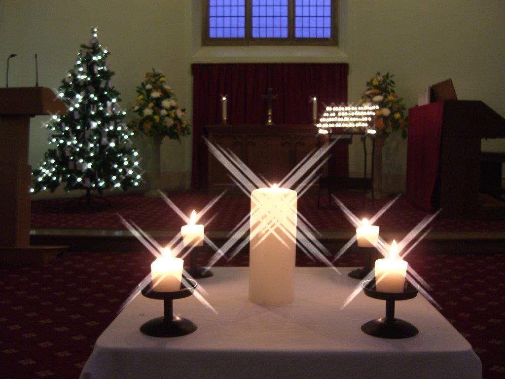 Five candles on a table in front of a Christmas tree. The candles are burning brightly and look like stars shining