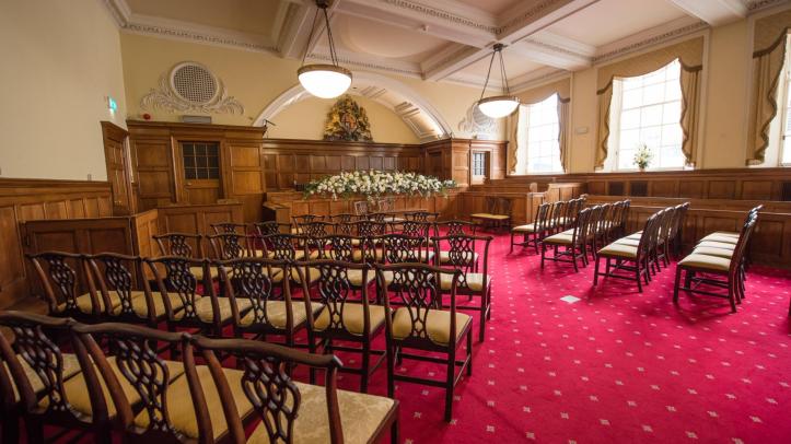 A wooden panelled room with a plush red carpet laid with wooden chairs arranged around a floral display