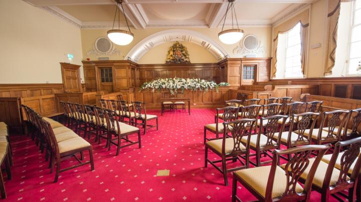 A wooden panelled room with a plush red carpet laid with wooden chairs arranged around a floral display