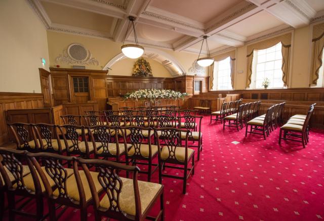 A wooden panelled room with a plush red carpet laid with wooden chairs arranged around a floral display