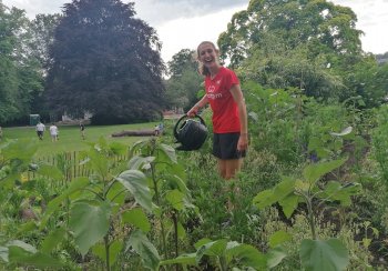 Woman from Good Gym waters the sunflowers with a watering can. She is wearing a red tshirt and smiling at the camera