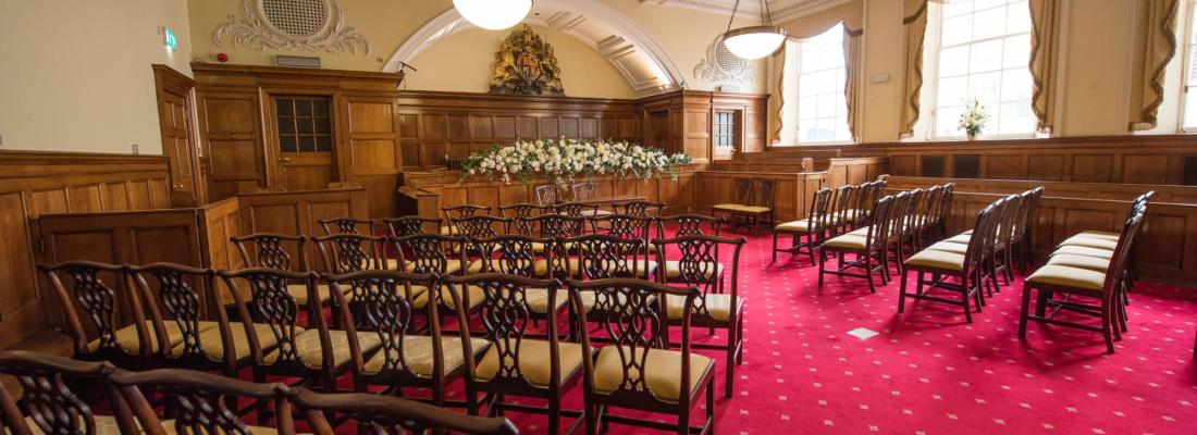 A wooden panelled room with a plush red carpet laid with wooden chairs arranged around a floral display